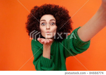 Positve friendly woman with Afro hairstyle in green casual style sweater making selfie POV, sending air kisses to followers while having livestream. Indoor studio shot isolated on orange background. 84888774