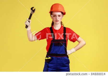 Portrait of confident builder woman holding adjustable wrench, looking at camera with serious expression, wearing overalls and helmet. Indoor studio shot isolated on yellow background. Portrait of confident builder woman holding adjustable wrench, looking at camera with serious expression, wearing overalls and helmet. Indoor studio shot isolated on yellow background. 84888814