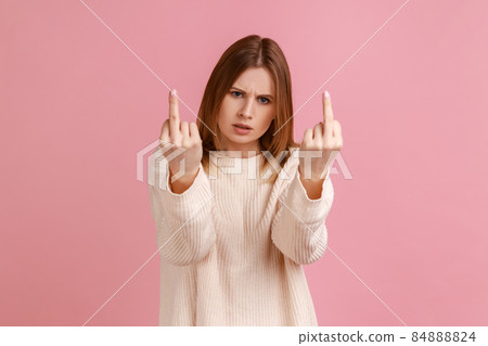 Portrait of blond woman rejecting communication, showing middle finger to express disrespect and hate, vulgar gesture, wearing white sweater. Indoor studio shot isolated on pink background. 84888824