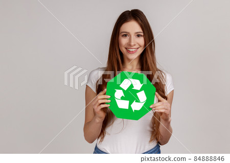 Portrait of beautiful positive woman holding recycling green symbol, sorting rubbish, saving environment, ecology, wearing white T-shirt. Indoor studio shot isolated on gray background. 84888846