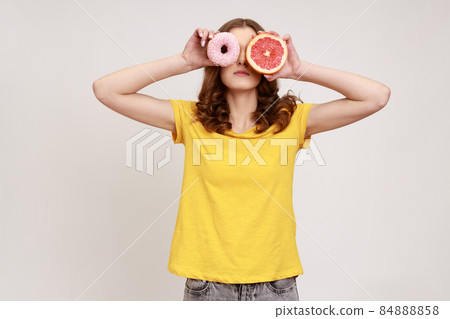 Sugary dessert vs fruits. Portrait of playful brown haired woman in yellow T-shirt covering eyes with fresh grapefruit and sweet doughnut, junk food. Indoor studio shot isolated on gray background. 84888858