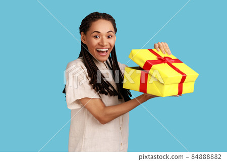 Portrait of happy amazed woman with black dreadlocks looking into gift box, opening present and looking at camera with happiness, wearing white shirt. Indoor studio shot isolated on blue background. Portrait of happy amazed woman with black dreadlocks looking into gift box, opening present and looking at camera with happiness, wearing white shirt. Indoor studio shot isolated on blue background. 84888882