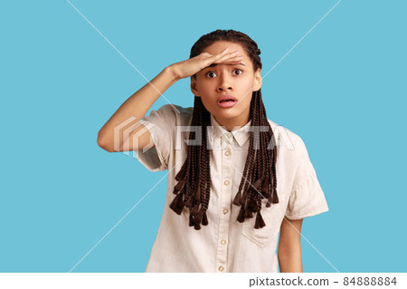 Young serious woman with black dreadlocks keeps hand near forehead, looks far away, searches something on horizon, wearing white shirt. Indoor studio shot isolated on blue background. 84888884