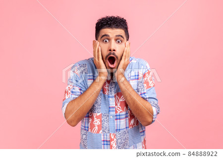 Portrait of astonished bearded man in blue shirt looking with big eyes, holding hands on face and screaming in amazement, shocked by sudden event. Indoor studio shot isolated on pink background. 84888922