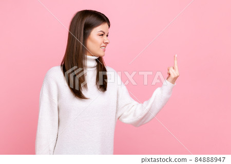 Side view portrait of aggressive woman standing and shows fuck sign, expressing negative emotions, wearing white casual style sweater. Indoor studio shot isolated on pink background. 84888947