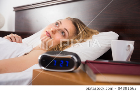 woman lies on bed with eyes open in bedroom near clock and book woman lies on bed with eyes open in bedroom near clock and book 84891662