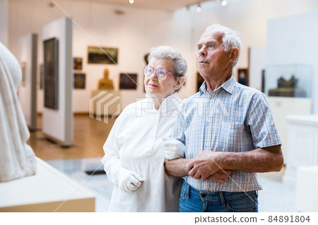 mature European couple examines paintings in an exhibition in hall of art museum 84891804