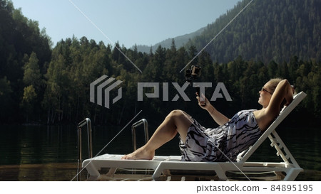 Woman shooting on handheld film gimbal stabilization for smartphone. Girl lie sunbed on pier make selfie. Blogger broadcast video vlogging. 84895195