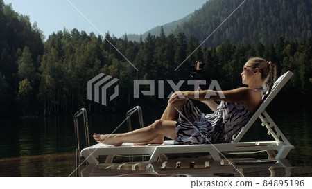 Woman shooting on handheld film gimbal stabilization for smartphone. Girl lie sunbed on pier make selfie. Blogger broadcast video vlogging. 84895196