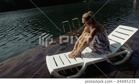 Woman lie on a sunbed in sunglasses and a boho silk shawl. Girl rest on a flood wood underwater pier. The pavement is covered with water in the lake. 84895235