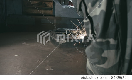 Forge workshop. Smithy. Worker in a welding hood helmet welds a part by electric welding. Sparks are reflected in the protective screen. 84896203