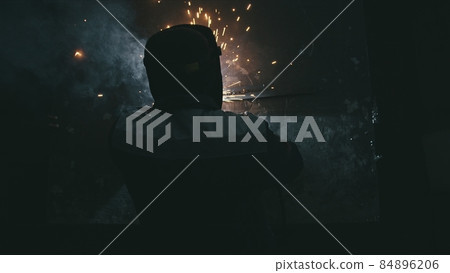 Forge workshop. Smithy. Worker in a welding hood helmet welds a part by electric welding. Sparks are reflected in the protective screen. 84896206