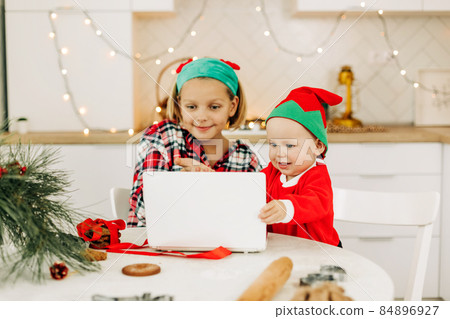 Brother and sister are sitting in the kitchen and congratulating their grandparents on Christmas and New Year via video link. Remote celebration of the online Christmas holiday Brother and sister are sitting in the kitchen and congratulating their grandparents on Christmas and New Year via video link. Remote celebration of the online Christmas holiday 84896927