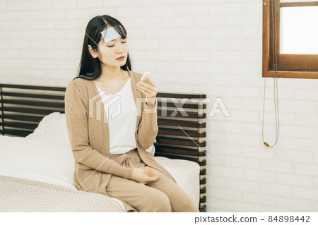 一位健康狀況不佳的女性,穿著涼爽的床單和睡衣坐在床上,用溫度計測量體溫 一位健康狀況不佳的女性,穿著涼爽的床單和睡衣坐在床上,用溫度計測量體溫 84898442