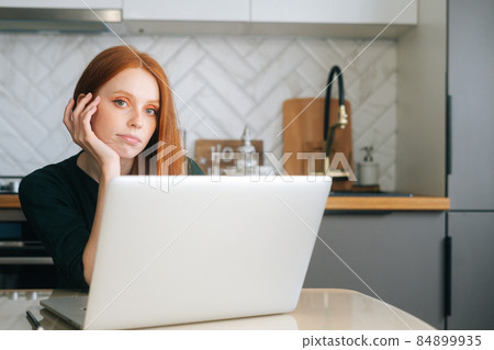 Medium shot of tired young woman typing on laptop keyboard holding hand on chin sitting, looking at camera. Medium shot of tired young woman typing on laptop keyboard holding hand on chin sitting, looking at camera. 84899935