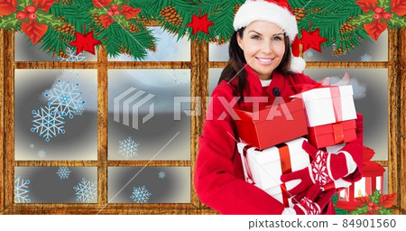 Composition of happy woman in santa hat carrying gifts against decorated window, copy space 84901560