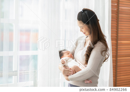Beautiful Asian woman holding newborn baby in her arms standing in front of windows at cozy home.Happy infant baby sleep in mother arms safety and comfortable.Mom and Baby Concept Beautiful Asian woman holding newborn baby in her arms standing in front of windows at cozy home.Happy infant baby sleep in mother arms safety and comfortable.Mom and Baby Concept 84902266