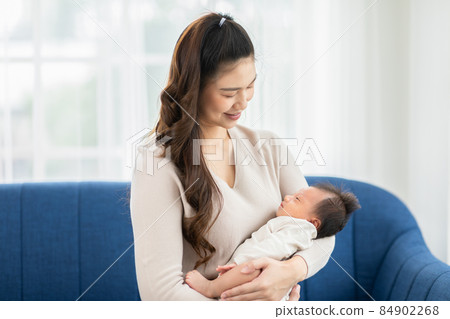 Beautiful Asian woman holding newborn baby in her arms sit on couch at white cozy home.Happy infant baby sleep in mother arms safety and comfortable.Mom and Baby Concept 84902268