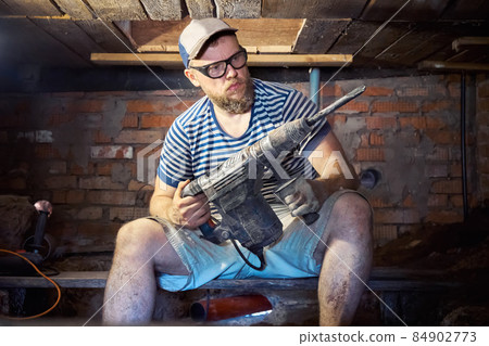 Funny bearded man with heavy duty perforator in the basement of the house 84902773