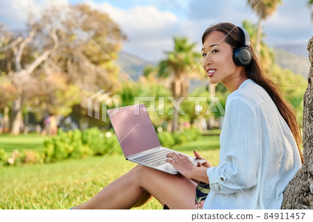 Young smiling woman in headphones using laptop sitting in park on grass Young smiling woman in headphones using laptop sitting in park on grass 84911457