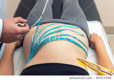 Male physiotherapist applying blue adhesive elastic therapeutic tape on female abdominal muscle of young adult slim sporty woman lying on couch doctor office. Injury rehabilitation and healthcare 84915496