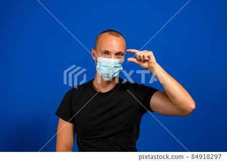 I need a few more. Portrait of young man with surgical medical mask in black casual style shirt standing and looking at camera and begging for something. Indoor studio shot, isolated blue background I need a few more. Portrait of young man with surgical medical mask in black casual style shirt standing and looking at camera and begging for something. Indoor studio shot, isolated blue background 84918297
