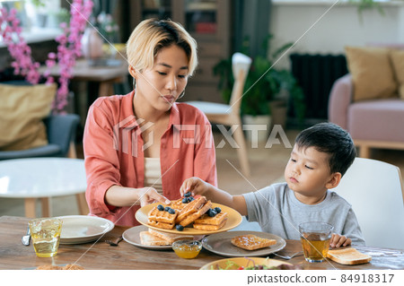 Young attractive woman passing her cute little son some waffles with blueberries Young attractive woman passing her cute little son some waffles with blueberries 84918317