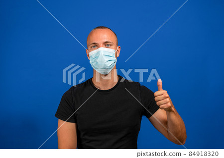 Like. Portrait of satisfied young worker man with surgical medical mask standing thumbs up and looking at camera smiling. indoor studio shoot isolated on blue background. Like. Portrait of satisfied young worker man with surgical medical mask standing thumbs up and looking at camera smiling. indoor studio shoot isolated on blue background. 84918320