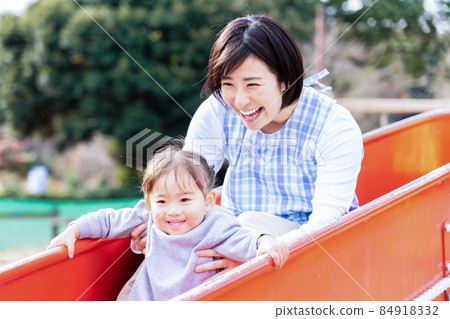 A 2-year-old girl and a young female nursery teacher playing on a slide A 2-year-old girl and a young female nursery teacher playing on a slide 84918332