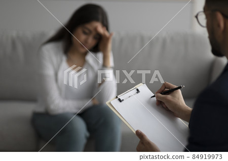 Depressed middle eastern lady consulting psychologist at medical office, focus on psychiatrist writing in clipboard Depressed middle eastern lady consulting psychologist at medical office, focus on psychiatrist writing in clipboard 84919973
