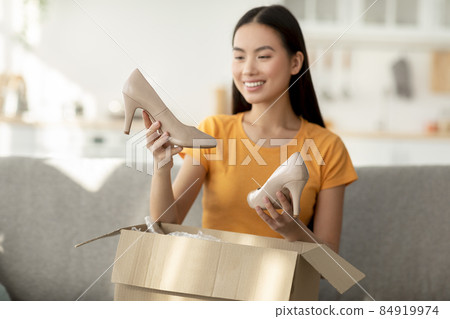 Satisfied asian woman unpacking cardboard box with new shoes, looking at high heels and smiling, sitting on sofa at home Satisfied asian woman unpacking cardboard box with new shoes, looking at high heels and smiling, sitting on sofa at home 84919974