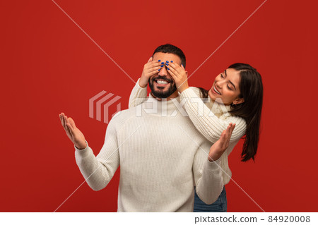 Romantic surprise concept. Cheerful arab woman covering her boyfriend eyes from back, red studio background 84920008