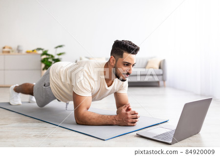 Online sports during coronavirus quarantine. Fit young Arab guy doing exercises near laptop at home 84920009
