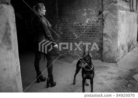 A girl in leather, with a Doberman, in an old yard. A girl in leather, with a Doberman, in an old yard. 84920070