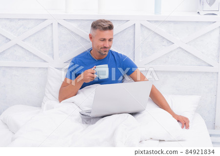 Awake from sleep. Awake man in bed. Happy guy drink tea using laptop. Morning relaxation 84921834