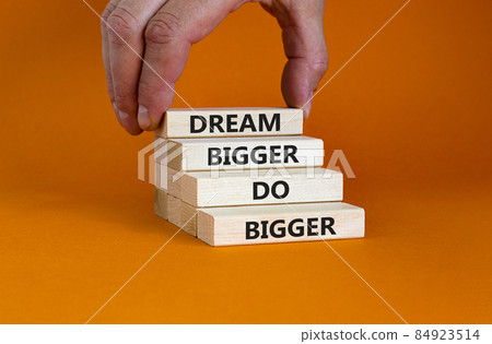 Dream and do bigger symbol. Concept words 'Dream bigger do bigger' on wooden blocks on a beautiful orange background. Businessman hand. Business, motivational and dream and do bigger concept. 84923514