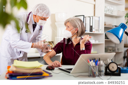 Focused doctor vaccinating aged businesswoman at work desk in office 84930251