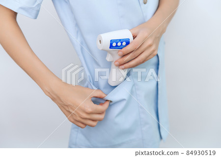 Temperature measurement gun in doctor hands. Close-up shot of doctor wearing protective surgical mask ready to use infrared isometric thermometer gun to check body temperature for virus symptoms. Temperature measurement gun in doctor hands. Close-up shot of doctor wearing protective surgical mask ready to use infrared isometric thermometer gun to check body temperature for virus symptoms. 84930519
