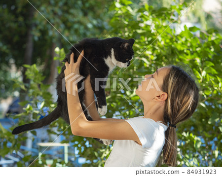 Teenager girl holds a black stray cat in her arms and looks at him. 84931923