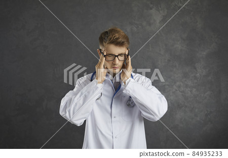 Medical work. Portrait of exhausted young male doctor experiencing severe headache and malaise. Sad doctor rubs his temples feeling tired from burning and insomnia standing on gray background. Medical work. Portrait of exhausted young male doctor experiencing severe headache and malaise. Sad doctor rubs his temples feeling tired from burning and insomnia standing on gray background. 84935233