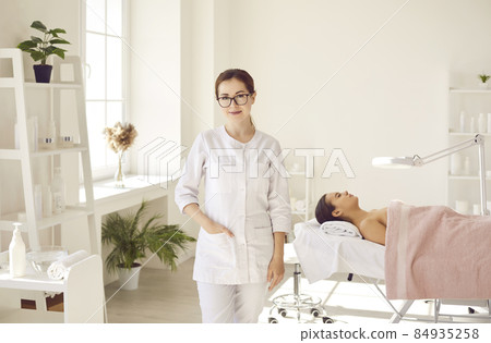 Female dermatologist, skin therapist, beautician and skincare professional with patient in office. Beautiful woman in coat uniform and glasses standing in room for examinations and cleaning procedures 84935258