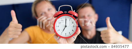 Young and happy man and woman holding red alarm clock and thumbs up while lying in bedroom Young and happy man and woman holding red alarm clock and thumbs up while lying in bedroom 84935477
