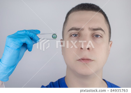 Conceptual shot of an eye crystalline lens replacement. Ophthalmic surgery. Return of sight. Removal of cataracts. Surgical intervention in the eyeball. Doctor holds the implant near the patient 84935763