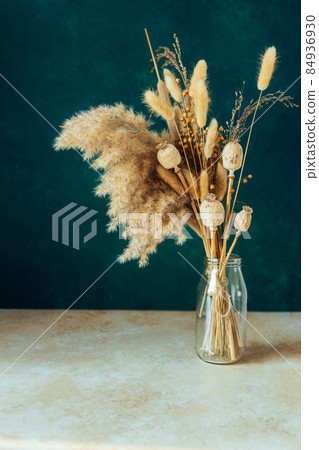 Bouquet of beige dried flowers in a glass vase on green-blue background. 84936930