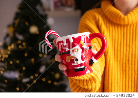 Woman unrecognizable in orange sweater hold santa claus mug with sugar cane, christmas tree background.  84938824