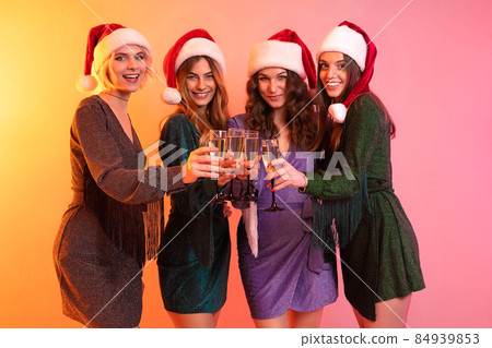 Four cheerful girls in Santa Claus hats holds glasses champagne having fun. New Year party Four cheerful girls in Santa Claus hats holds glasses champagne having fun. New Year party 84939853