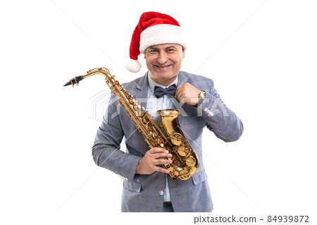 Funny musician wears in Santa's hat holds saxophone while straightening bow tie on studio background Funny musician wears in Santa's hat holds saxophone while straightening bow tie on studio background 84939872