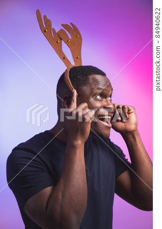 Portrait of african american man wears rudolph deer horns enjoy his accessory for christmas eve party celebration on colourful background Portrait of african american man wears rudolph deer horns enjoy his accessory for christmas eve party celebration on colourful background 84940622
