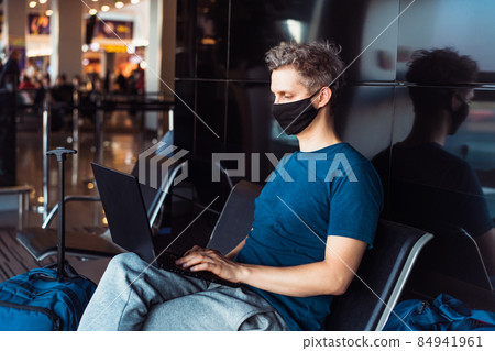 Young man wearing face mask and using laptop for remote work while sitting with distance at airport. New normal, digital nomad, freelance lifestyle. Traveling during pandemic, personal protection Young man wearing face mask and using laptop for remote work while sitting with distance at airport. New normal, digital nomad, freelance lifestyle. Traveling during pandemic, personal protection 84941961