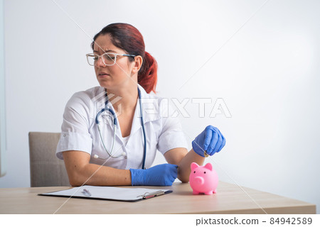 An upset female doctor puts a coin in a piggy bank on a white background. An upset female doctor puts a coin in a piggy bank on a white background. 84942589